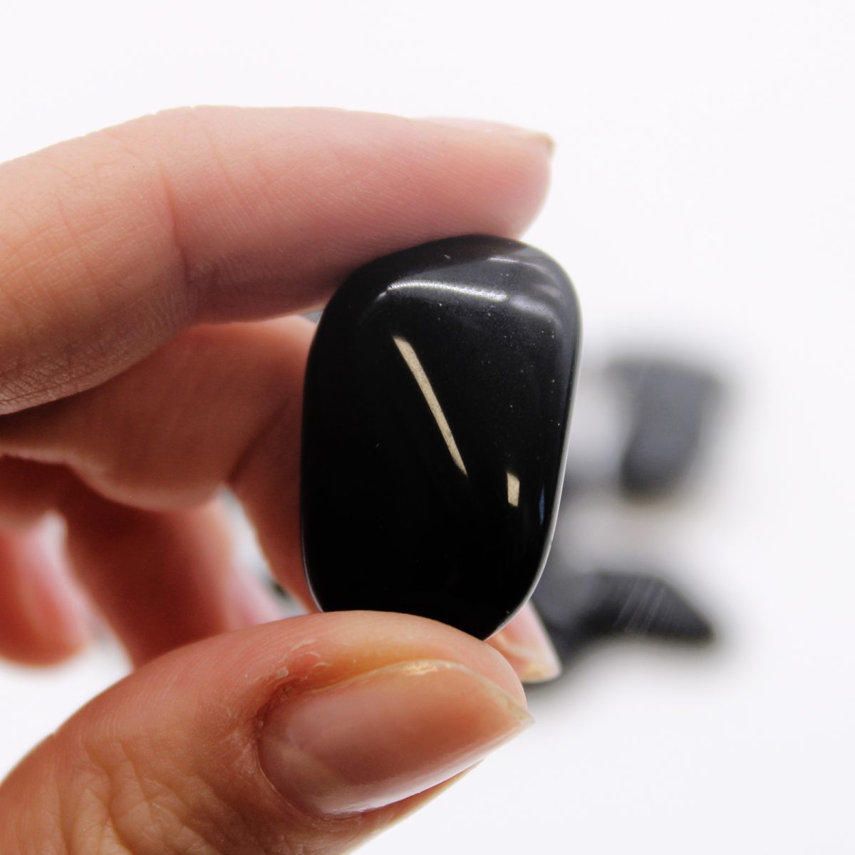 A hand holds a polished, oval, black stone between its fingers. More of the stones sit blurred in the white background.