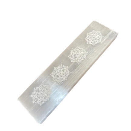 A selenite crystal bar rests on a white surface, adorned with four repeating mandala patterns, adding a decorative element to the crystal's natural striated texture.