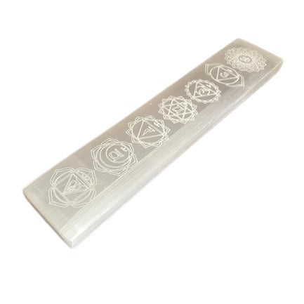 A selenite wand displays the seven chakra symbols linearly along its length. The rectangular wand is placed on a clean, white surface.
