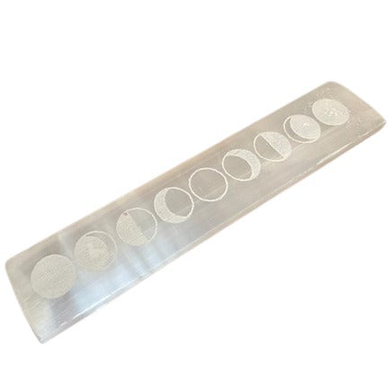 A rectangular, translucent selenite bar displays engraved phases of the moon in a row. It is diagonally oriented on a bright, white background.