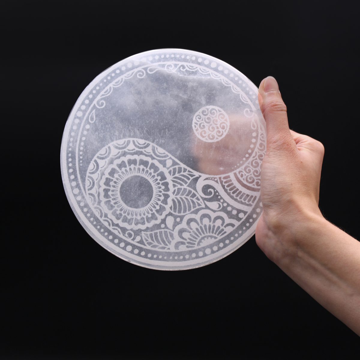 A hand holds a circular, translucent yin-yang symbol, delicately etched with floral designs and dots, against a solid black background.