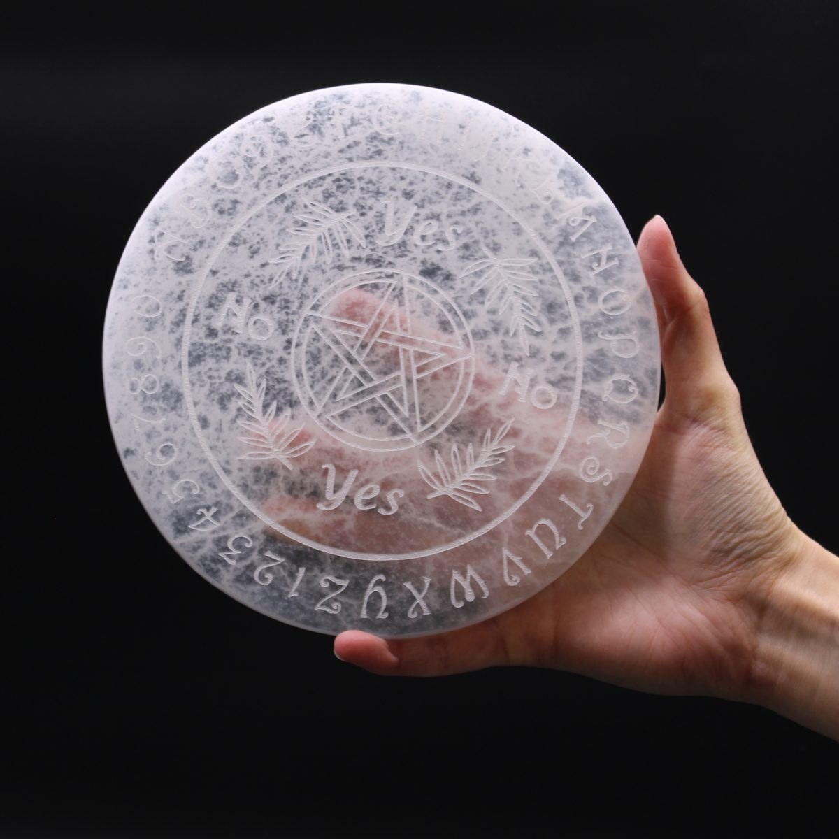 A Ouija board featuring letters, numbers, "Yes," and "No," is being held in a person's hand. The background is a plain, dark color.