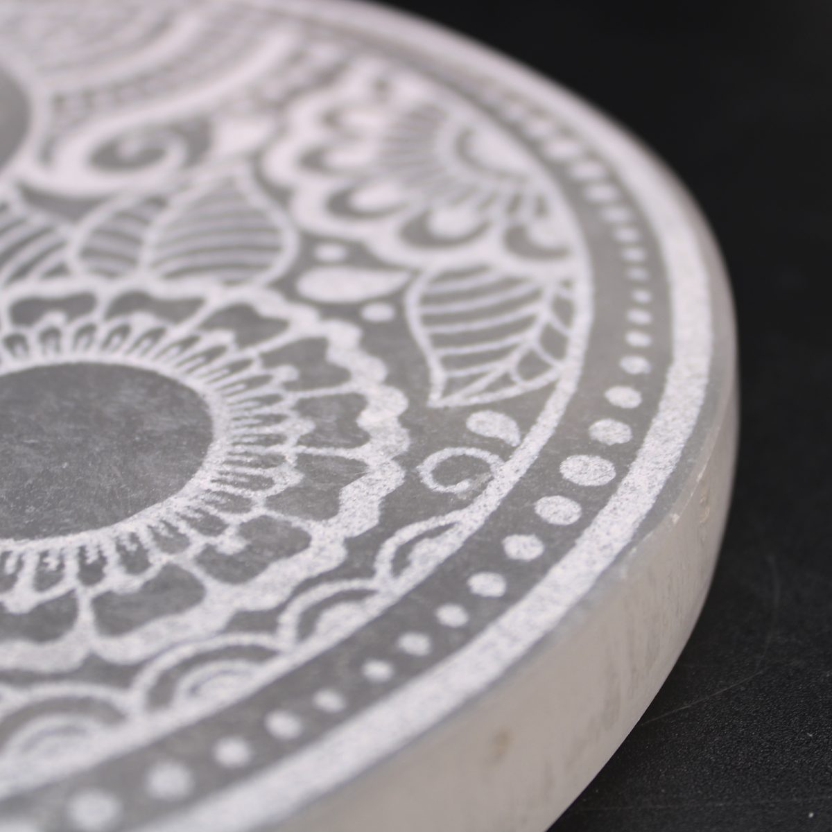 A round, white object features a detailed, floral design. The piece is positioned on a black surface, with the pattern’s delicate lines creating an intricate aesthetic.