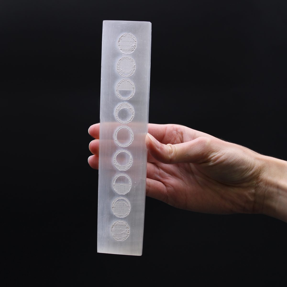 A hand holds a white, rectangular bar etched with a vertical series of circles. Each circle contains a different amount of shading within, mimicking lunar phases. The background is solid black.