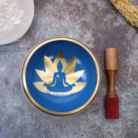 A blue singing bowl with a gold silhouette of a meditating figure in a lotus motif, placed on a textured surface beside a wooden mallet with a red felt end.