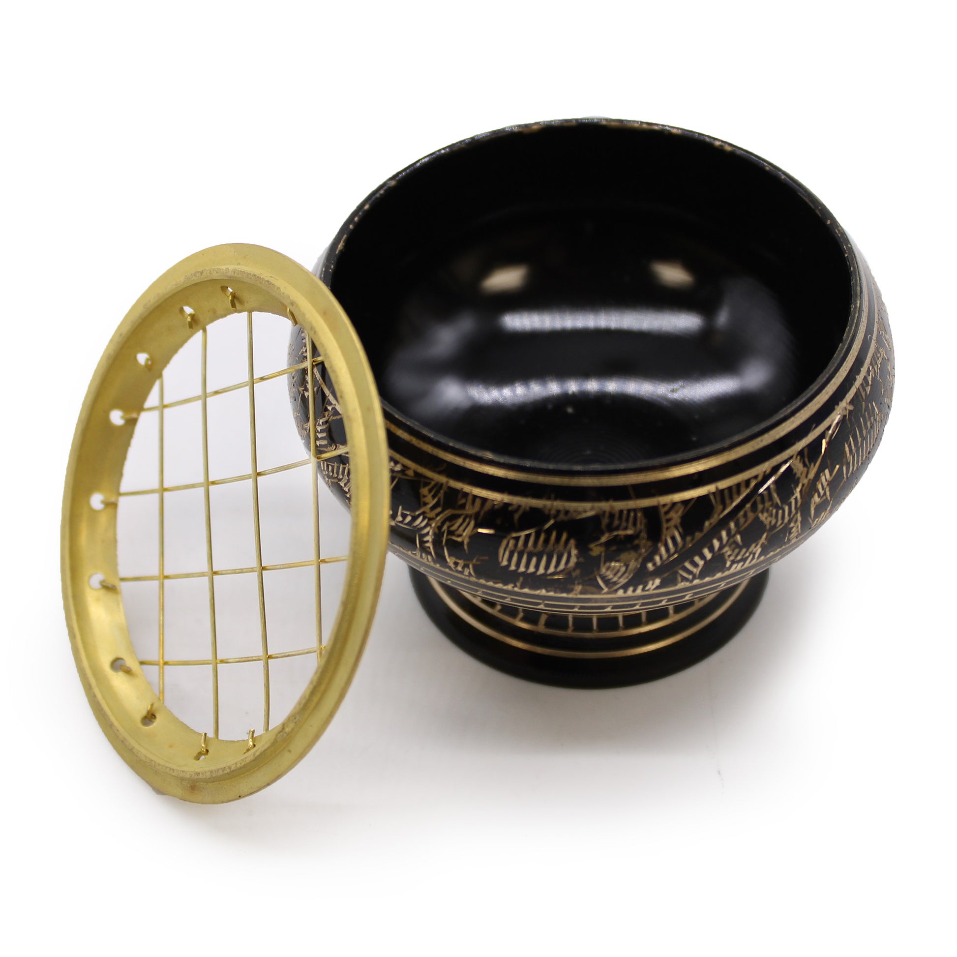 A black and gold incense burner with intricate carvings sits on a flat surface. A matching round metal grate, with a grid pattern, is placed beside it.