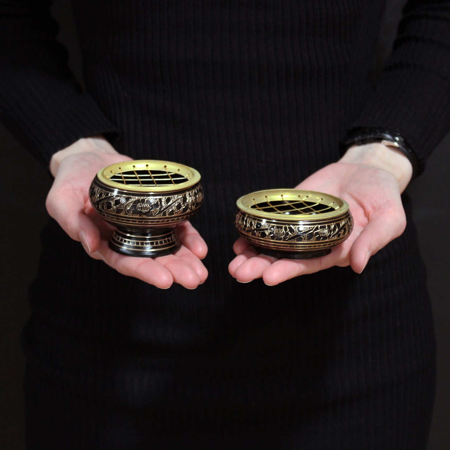 Two intricately designed brass incense burners, with decorative patterns, are held in hands against a dark background. The burners feature a wire mesh on top for burning incense safely.