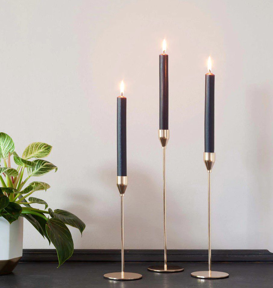 Three black candles burn atop golden candlesticks. The candles and a potted plant are arranged on a dark surface in front of a pale wall.