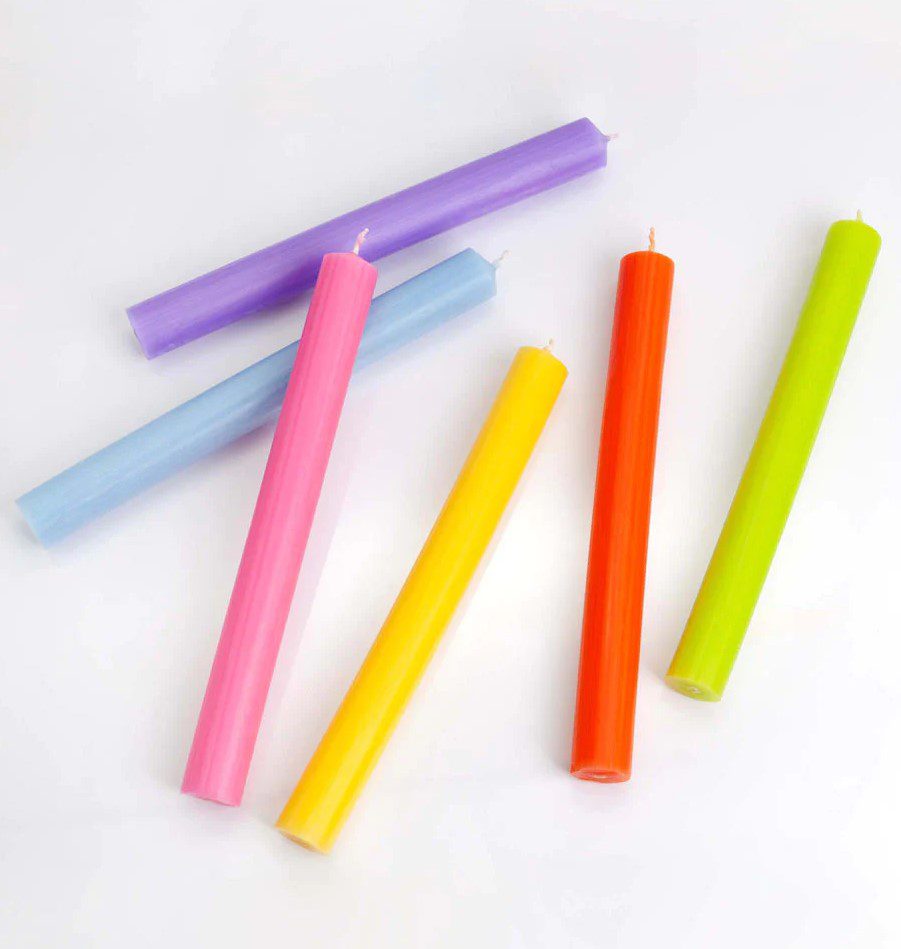 Six colorful, cylindrical candles lie on a plain white surface. The candles, each a different shade, are scattered irregularly.