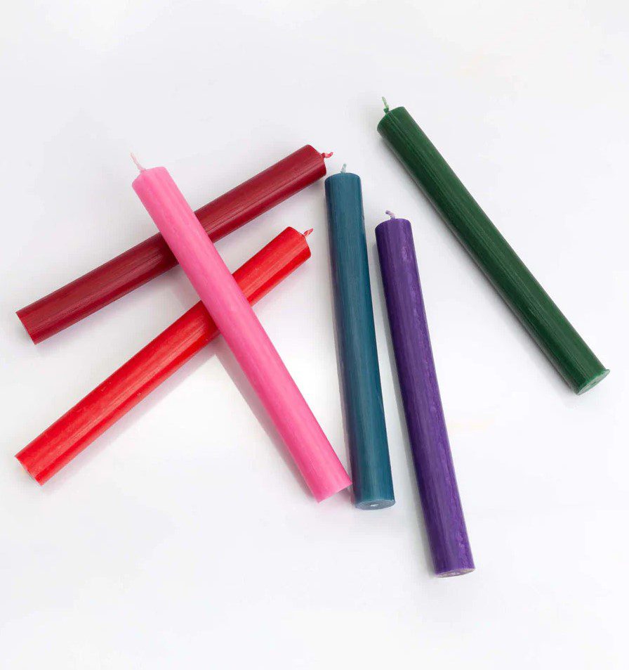 Several cylindrical candles of varying colors lie on a white surface. The colors include pink, red, burgundy, green, blue, and purple.