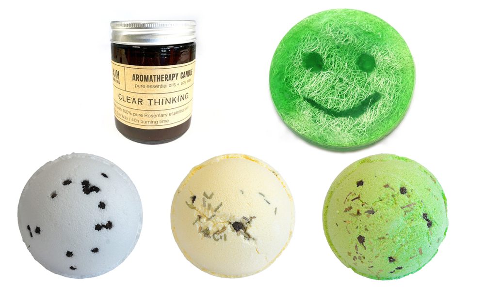 A brown aromatherapy candle, labeled "CLEAR THINKING," sits beside three bath bombs and a green smiley-face bath soap.  All items are arranged on a white background.