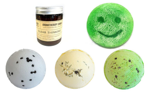 A brown aromatherapy candle, labeled "CLEAR THINKING," sits beside three bath bombs and a green smiley-face bath soap.  All items are arranged on a white background.