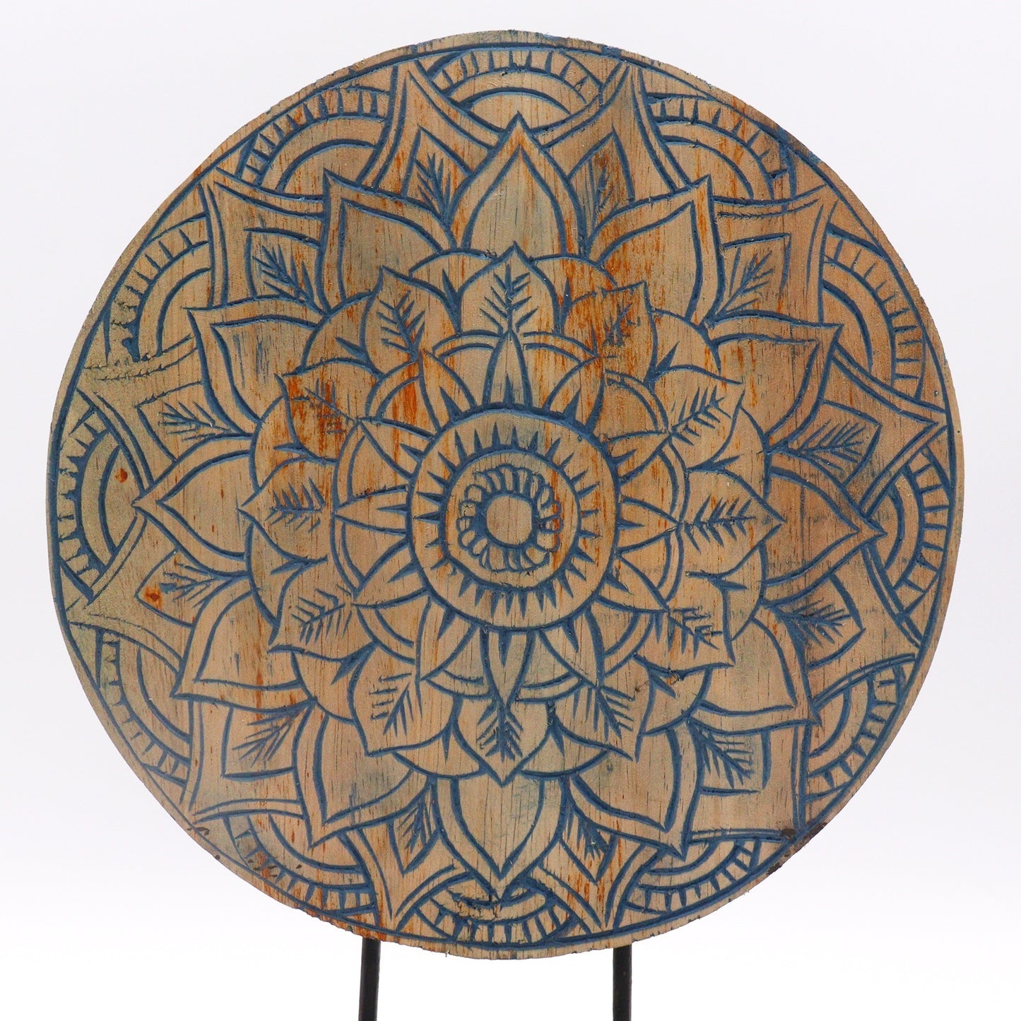 A round wooden disc displays an intricate, symmetrical mandala design with carved floral patterns, featuring a complex arrangement of petals and leaves highlighted with blue against a natural wood finish.