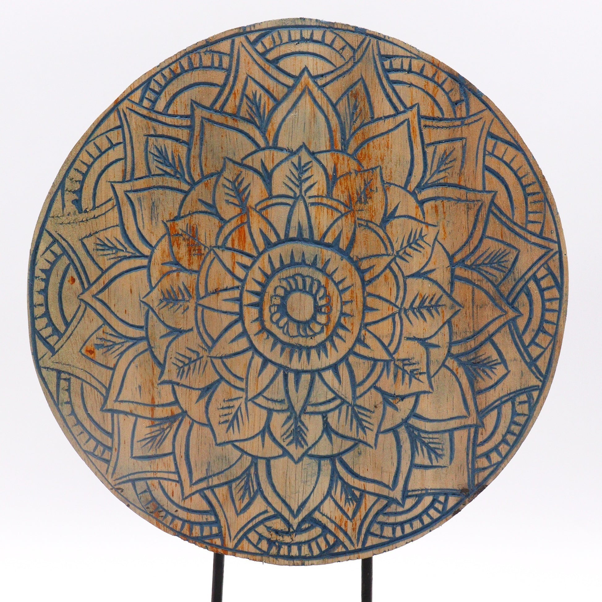 A round wooden disc displays an intricate, symmetrical mandala design with carved floral patterns, featuring a complex arrangement of petals and leaves highlighted with blue against a natural wood finish.
