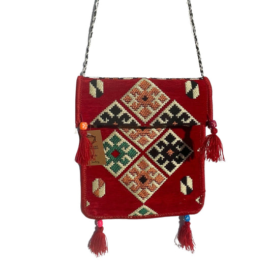 A red cross-body bag with diamond-patterned embroidery hangs against a white backdrop. The bag has a black and silver chain strap, tassels, and a tag that reads "ORIENT ROOM Handmade Turkey."