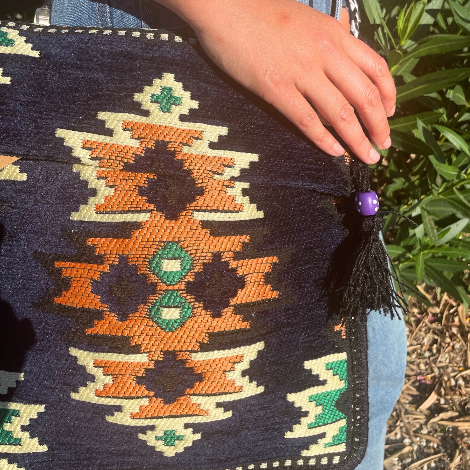 A navy patterned tapestry bag with orange, white, and green geometric designs is held by a person wearing denim jeans in an outdoor setting with green foliage. The bag has a black tassel with a purple bead.