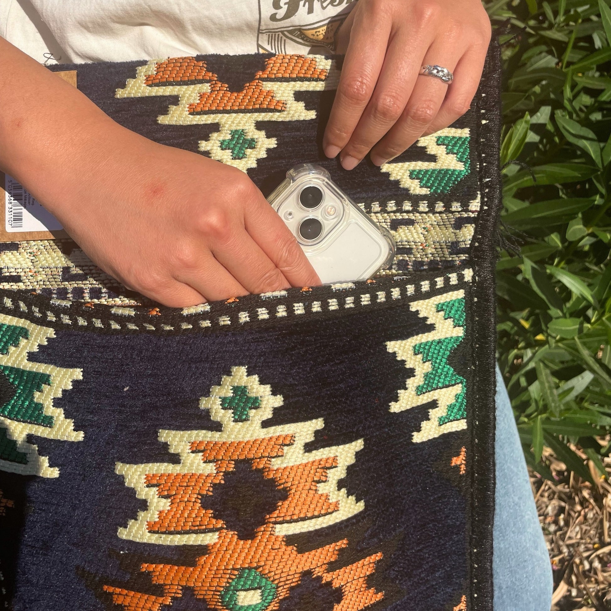 Hands place a smartphone in a clear case into a dark blue purse with an orange, white, and green southwestern pattern. The purse is held outdoors, in front of some greenery.