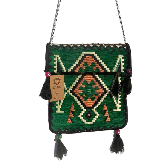 The green bag displays embroidered geometric patterns and hangs via a black and white rope strap. The tag reads "ANCIENT WISDOM Made In Turkey." Black tassels dangle with decorative beads.