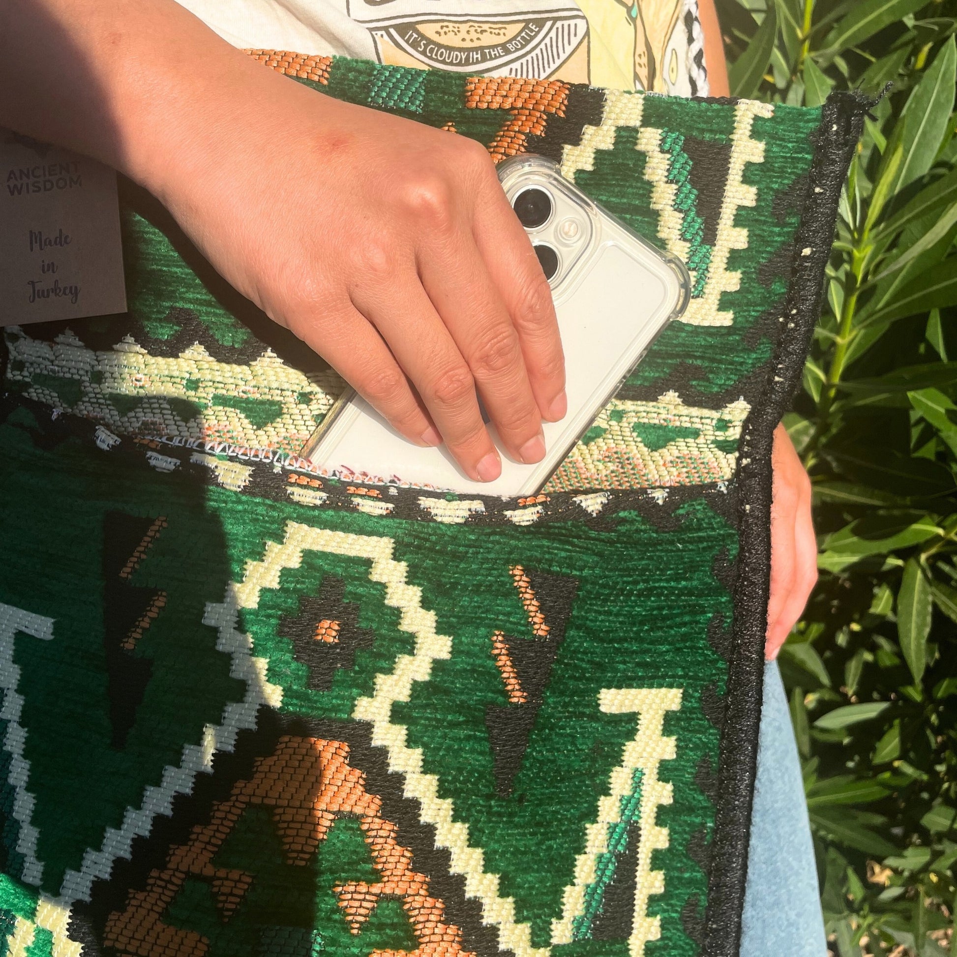 A hand inserts a clear-cased phone into the pocket of a green, patterned bag, partially obscuring the text "ANCIENT WISDOM, Made in Turkey". Text on the shirt reads "IT'S CLOUDY IN THE BOTTLE". Foliage is visible in the background.