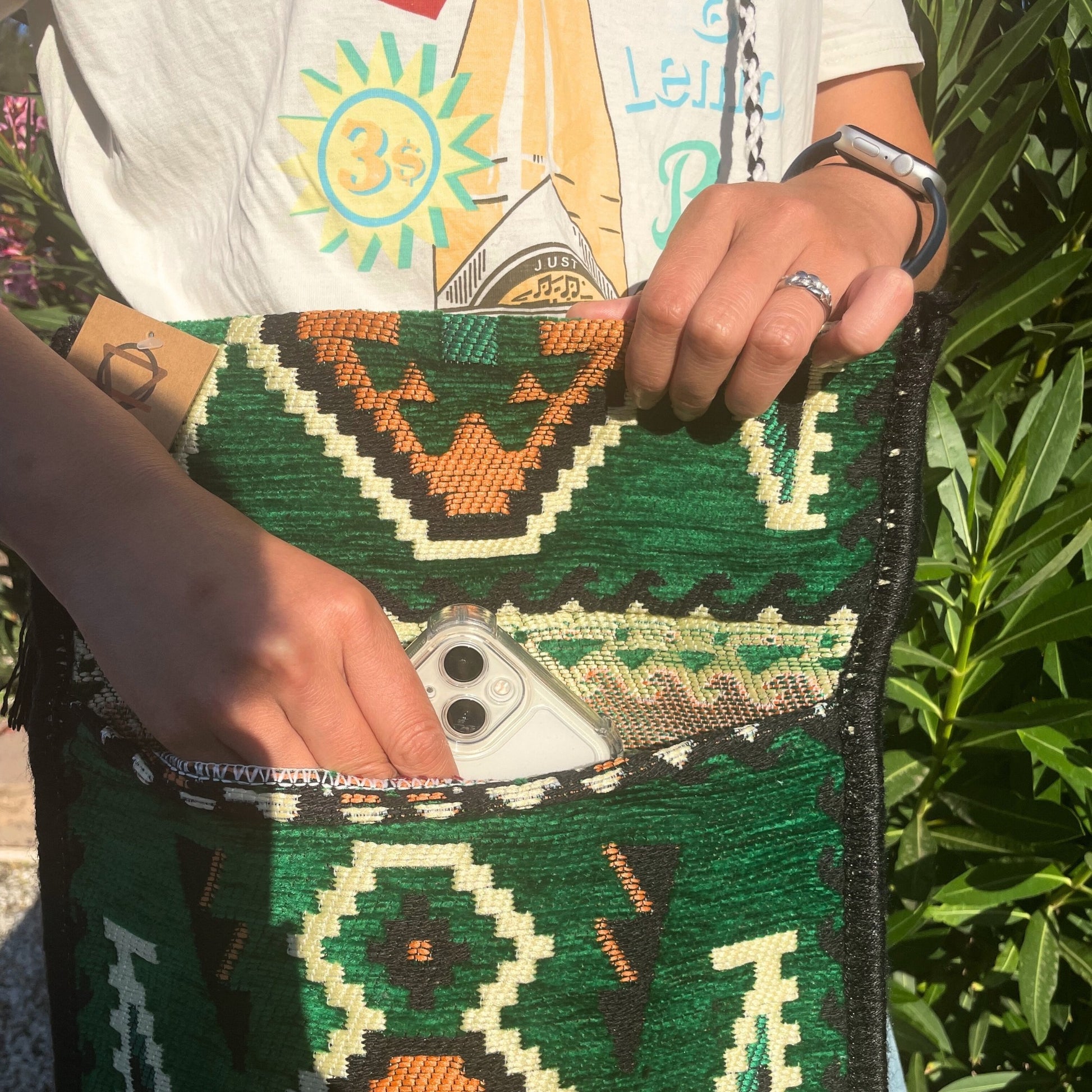 A person is putting a phone into a green, woven tote bag. The photo is taken outside near green foliage.