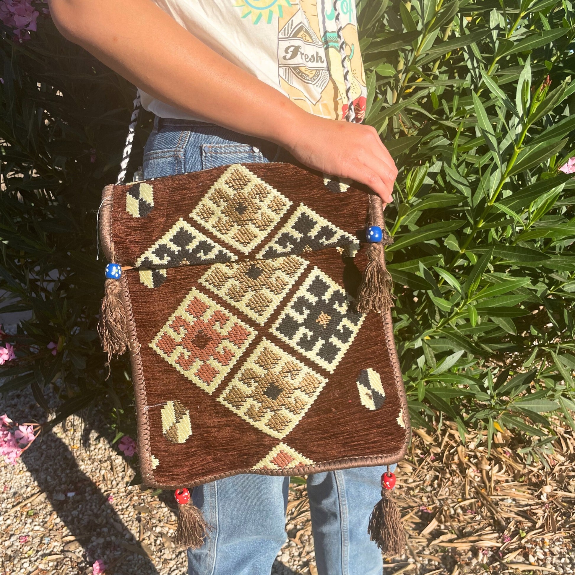 A person holds a brown, patterned shoulder bag with tassels and beads against a backdrop of vibrant greenery and pink flowers. The shirt reads, "Fresh IT'S CLOUDY BOTTLE."