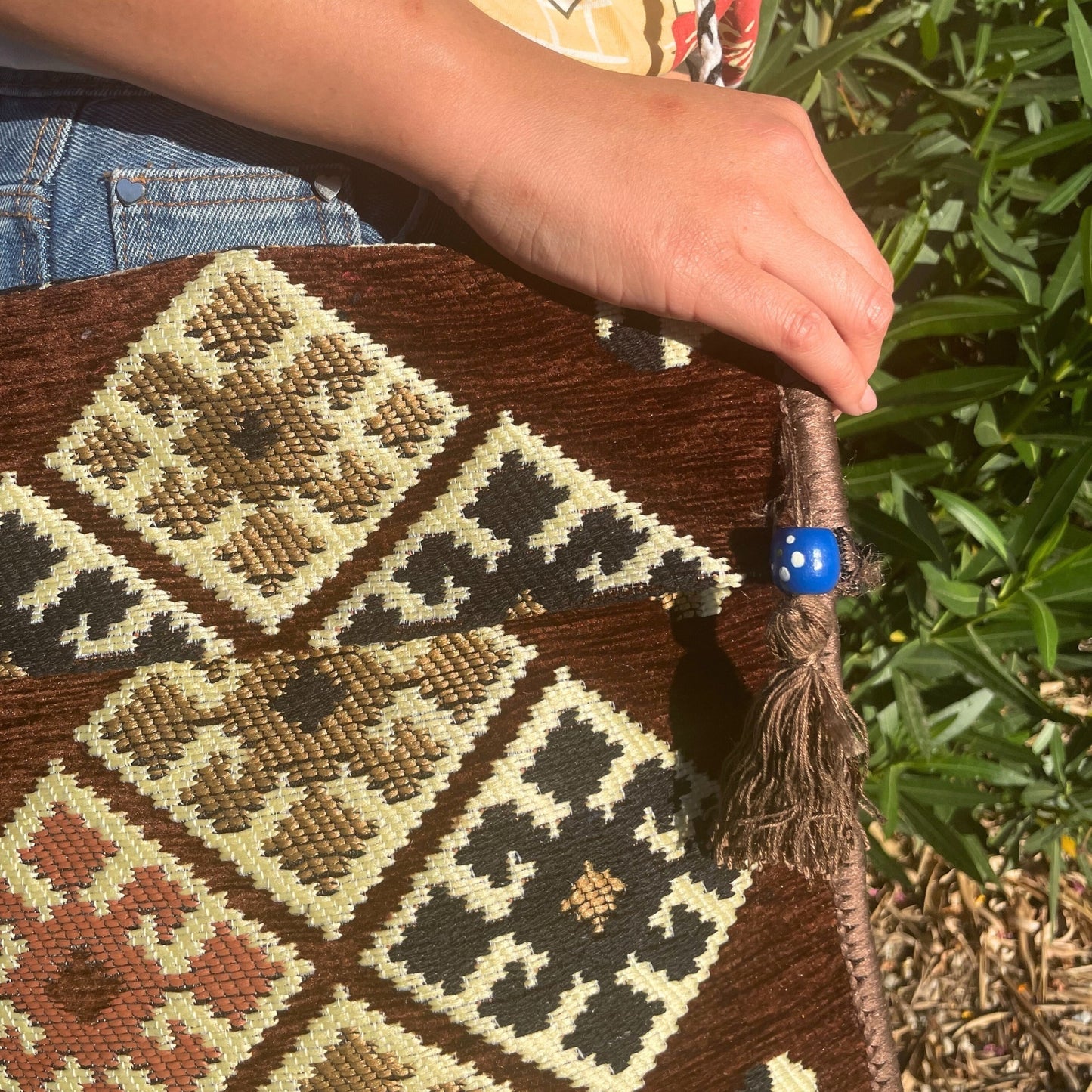 A hand rests on a textured, patterned brown tote bag held against blue jeans. A blue bead adorns a tassel on the bag, which sits in a sunny, grassy area.
