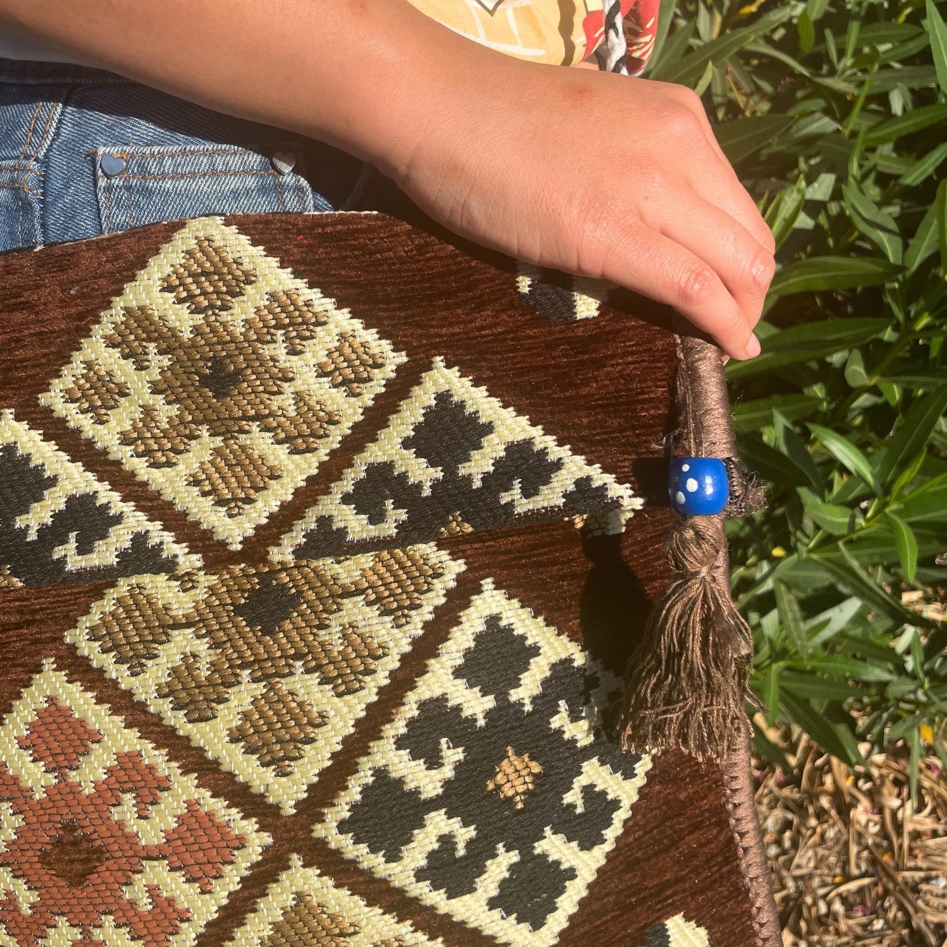 A hand rests on a textured, patterned brown tote bag held against blue jeans. A blue bead adorns a tassel on the bag, which sits in a sunny, grassy area.