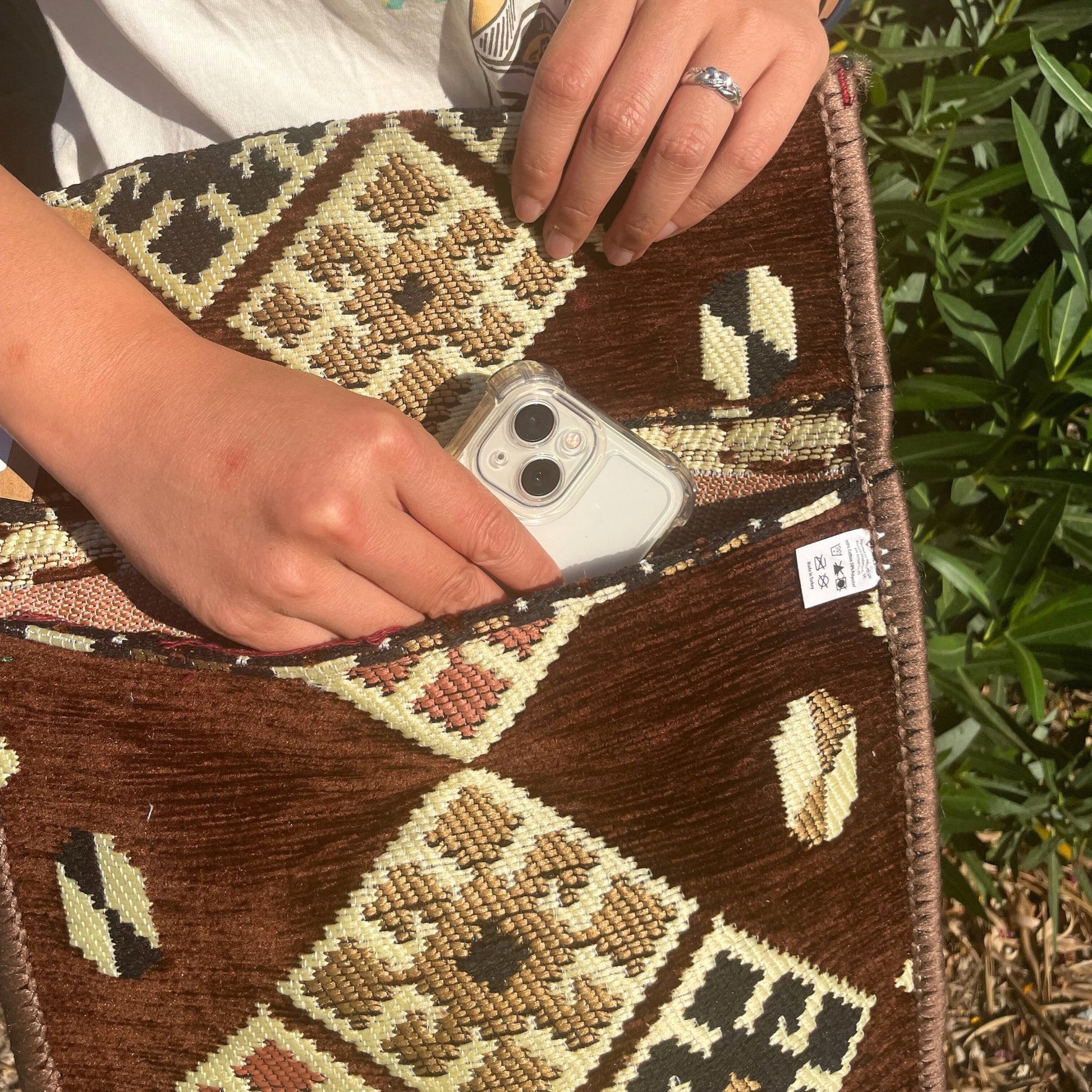 A person's hands place a phone into the pocket of a brown patterned textile bag. A white tag reads: "50% Cotton 50% Polyester Made in Turkey." The bag is outside amongst greenery.