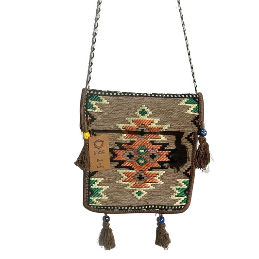 A woven purse with a brown strap hangs in a white space; it features geometric patterns and tassels. Tag reads: "ANCIENT WISDOM, Made in Turkey".