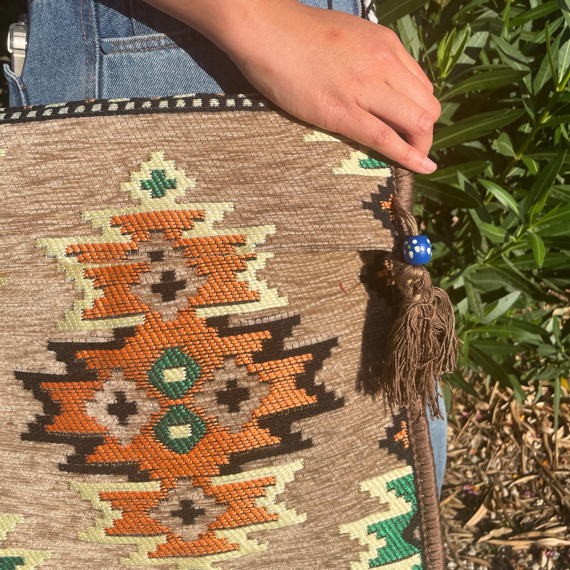 A patterned brown bag is held by a hand, with a blue beaded tassel visible. The background shows jeans and green foliage in natural light.
