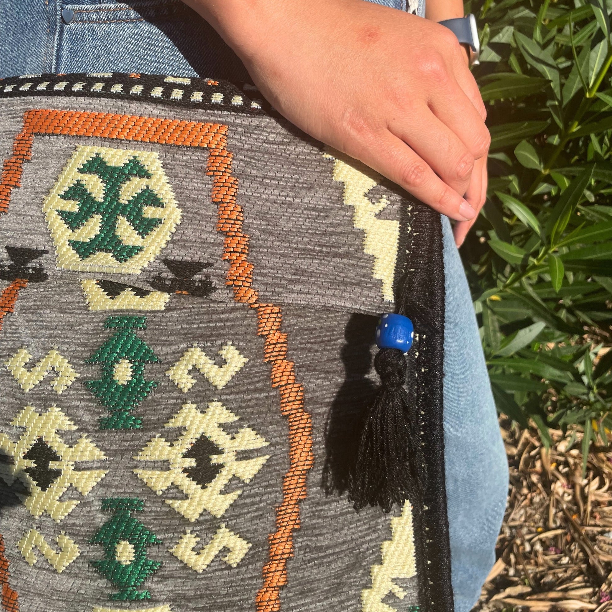 A person in jeans holds a gray patterned bag with orange, black, green, and cream accents, and a blue beaded tassel. Lush green bushes and wood chips are visible in the background.