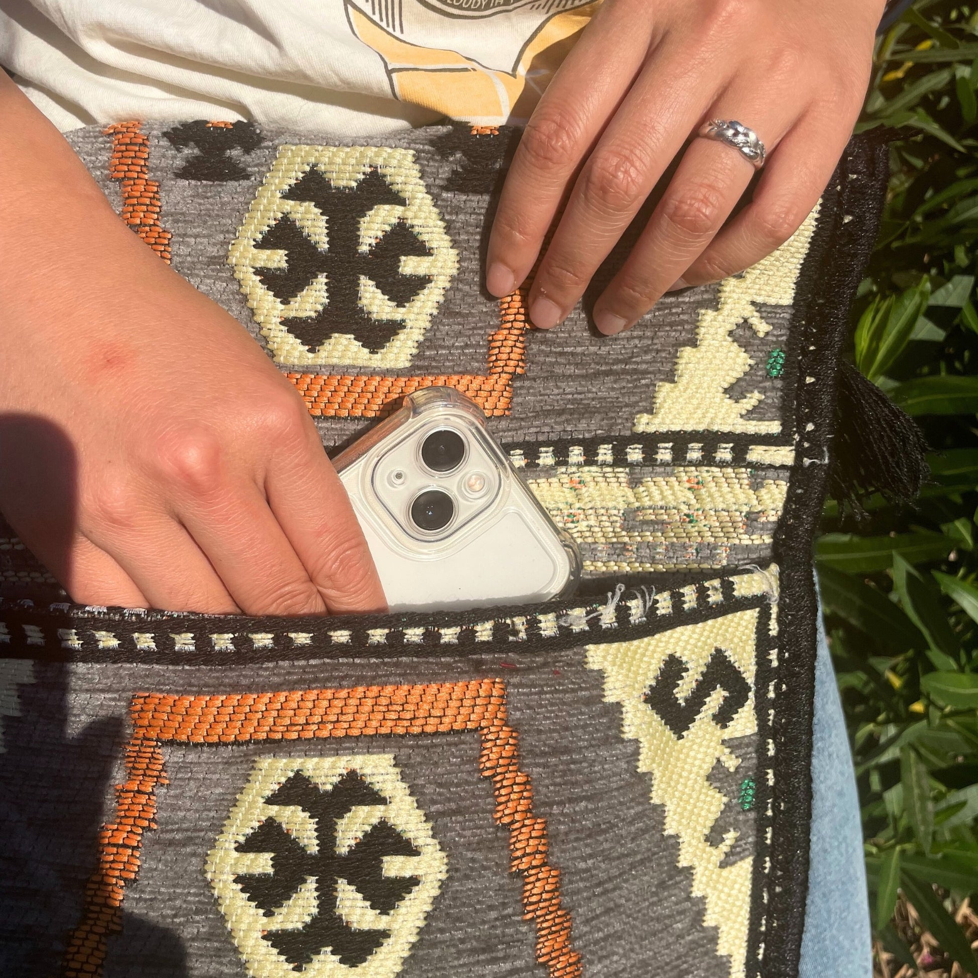 Hands place an iPhone in a woven bag with geometric patterns in yellow, black, and orange, while standing outside near greenery.