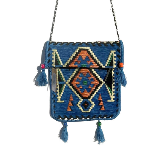 A blue woven purse hangs, adorned with geometric orange and green patterns. Tassels with colorful beads dangle along the purse's edges. The purse hangs against a white backdrop.