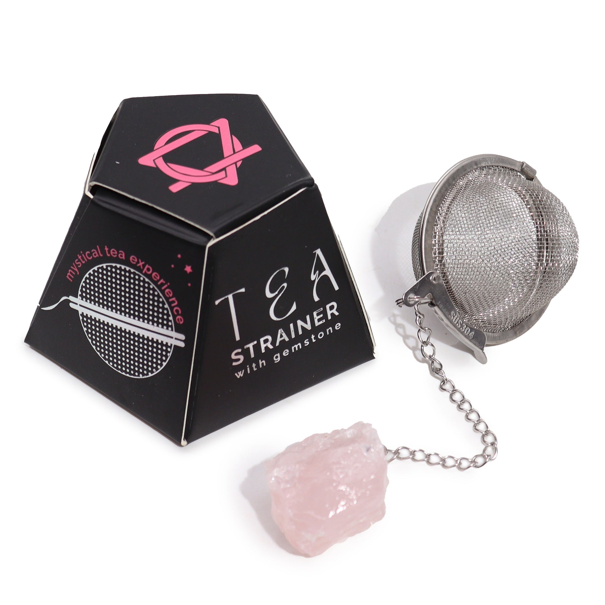 A metal tea strainer with a gemstone at the end of its chain lies next to black packaging labeled "TEA STRAINER with gemstone" and "mystical tea experience," featuring a pink design.