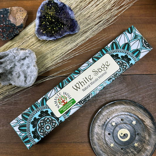 Incense package labeled "White Sage Natural Masala Incense" from Namaste India rests on a wooden surface with decorative crystals and a carved wood incense holder.