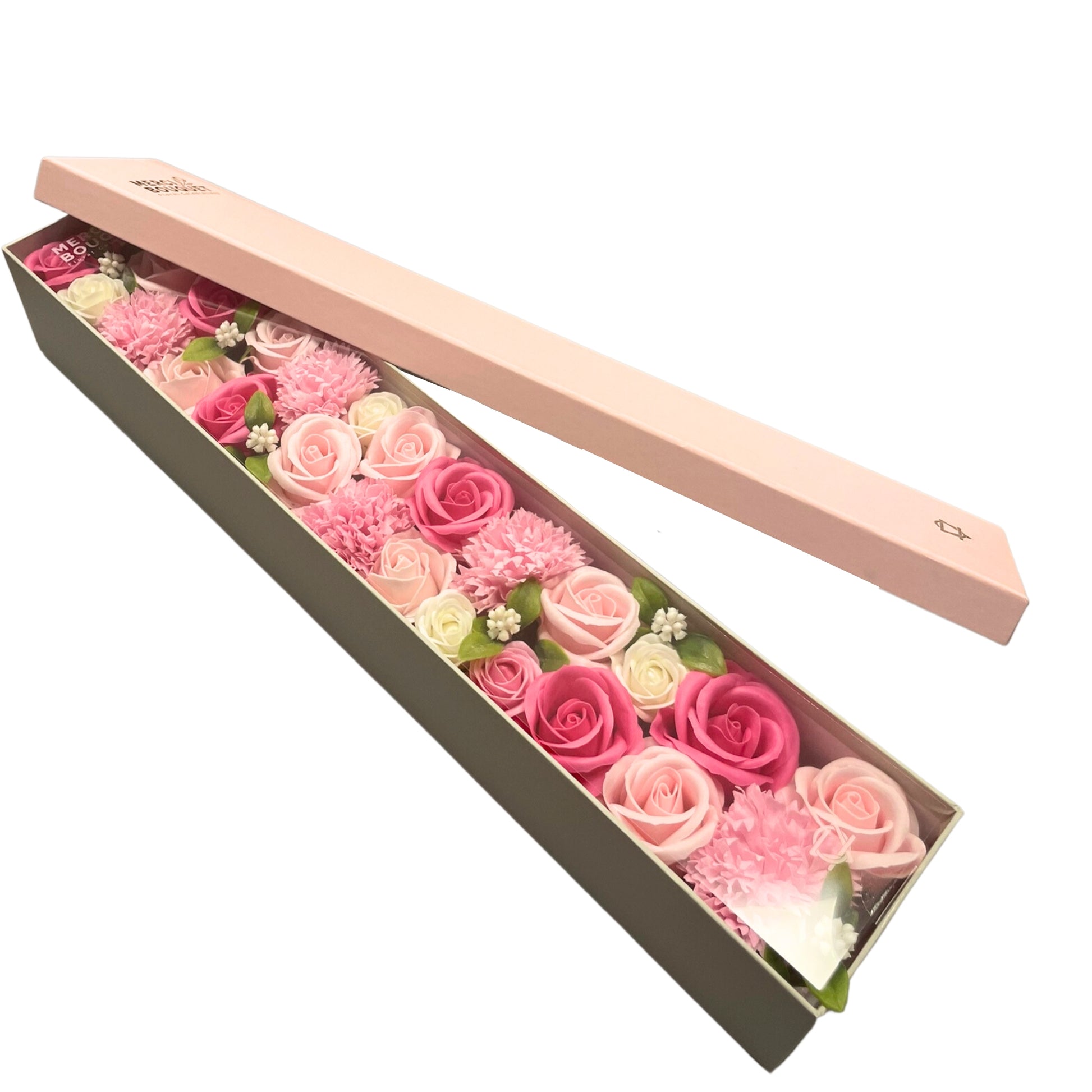 A long box containing soap flowers in shades of pink and white is open;  the lid rests beside it.  The flowers are arranged densely. MERCI BOUQUET is printed on the box lid.