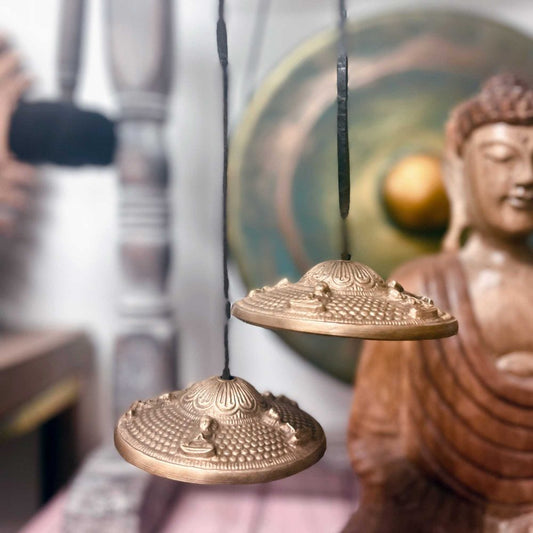 Bronze Tibetan cymbals hanging by cords, detailed with Buddha figures, are in front of a blurred Buddha statue and a colorful, circular backdrop, suggesting a serene, spiritual setting.