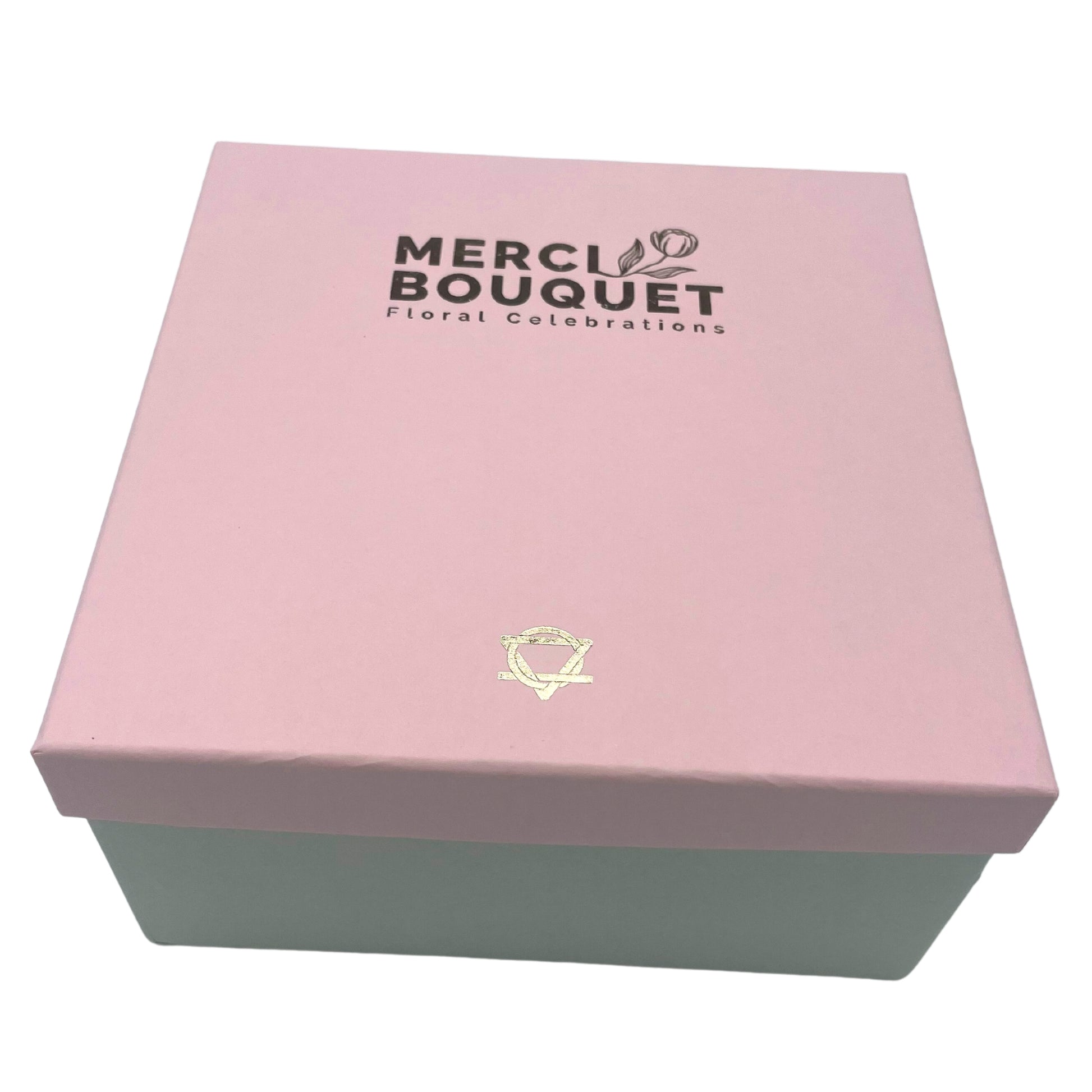 A pink box with a light green base sits on a surface.  The lid displays "MERCI BOUQUET Floral Celebrations".  A gold symbol is centered below the text.
