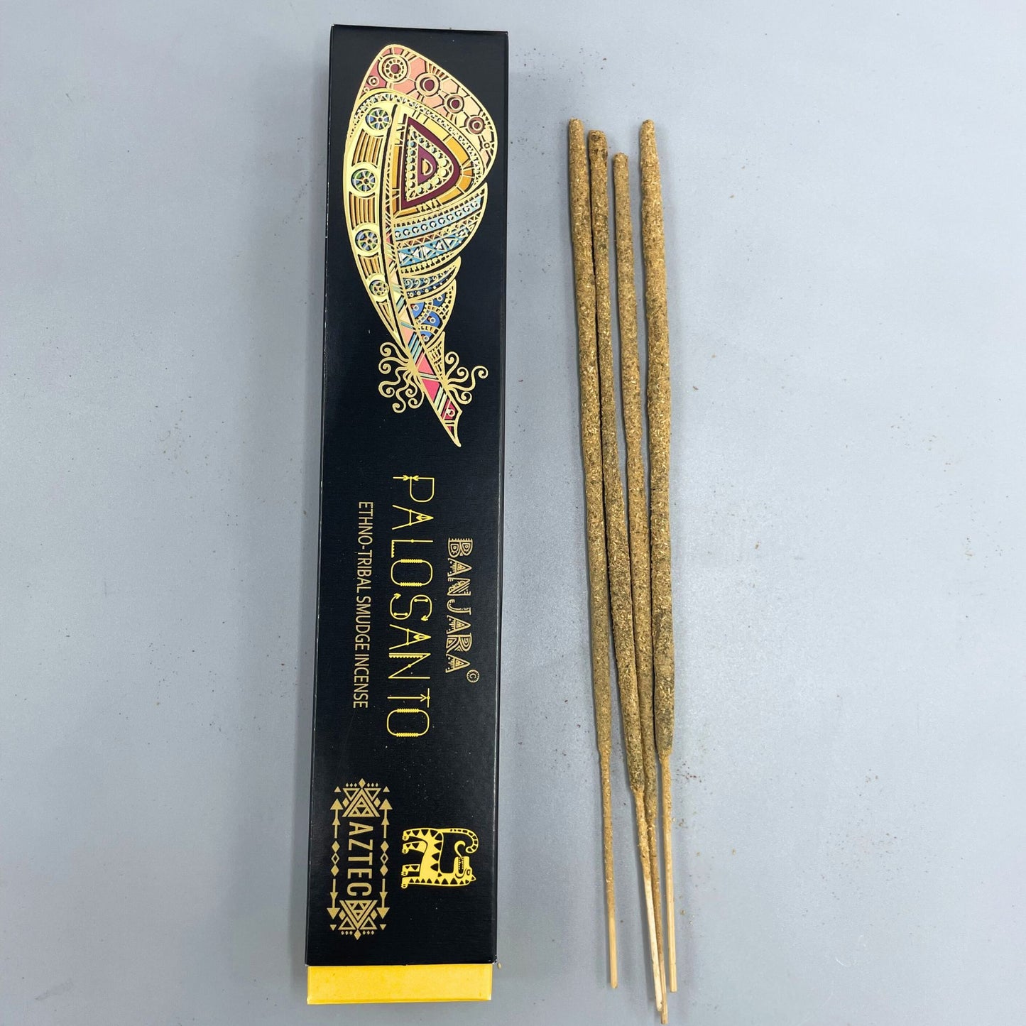 Box labeled "BANJARA® PALOSANTO Ethno Tribal Smudge Incense," featuring a colorful feather design, lies next to four incense sticks on a light gray surface. "AZTEC" is printed with tribal motifs.