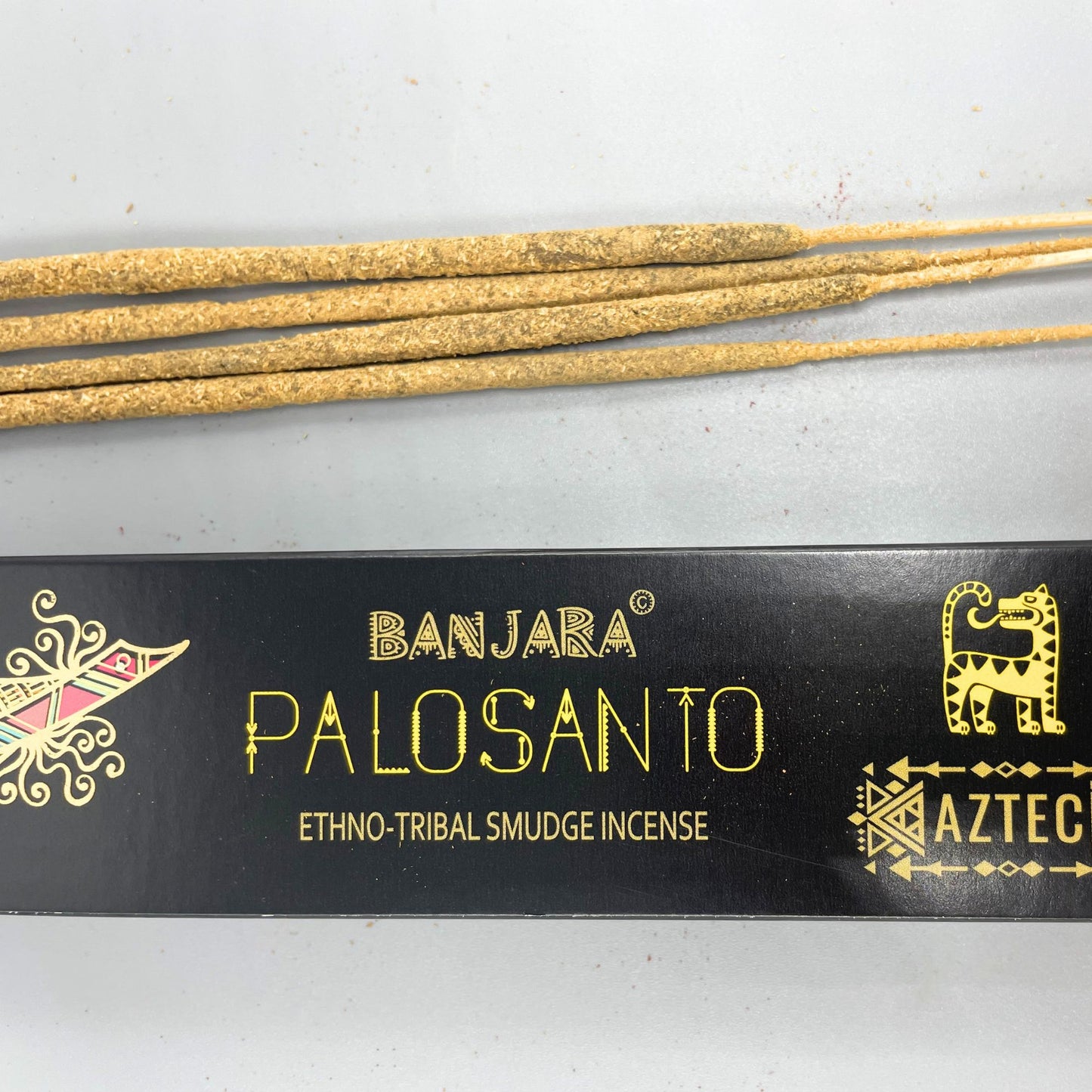 Incense sticks lie on a light surface. Below them, a black box labeled "BANJARA PALO SANTO ETHNO-TRIBAL SMUDGE INCENSE AZTEC" features tribal designs and a stylized animal.
