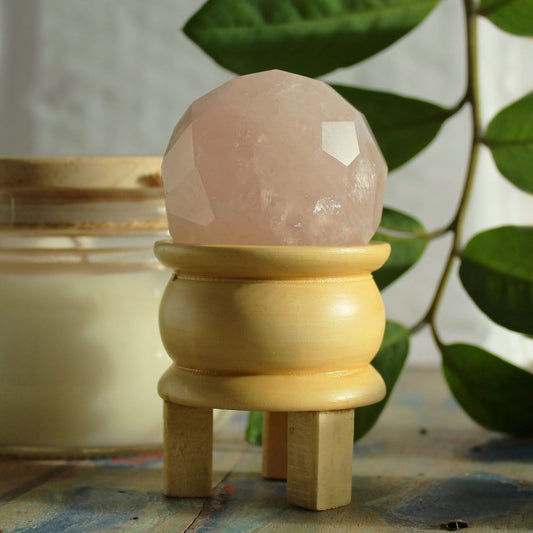 A faceted rose quartz sphere rests on a small, light-wooden stand.  The stand has three legs.  The setting includes a candle and leafy greenery.