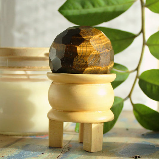 A faceted tiger eye sphere rests on a small, light-colored wooden stand.  The stand has four legs. It's situated near a candle and leafy plant.