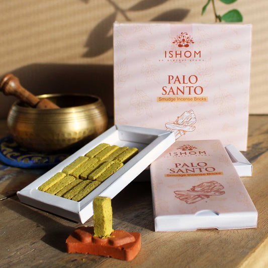Incense bricks displayed in an open box titled "ISHOM Palo Santo Smudge Incense Bricks." A single brick stands upright on a clay holder. A brass bowl is in the background.