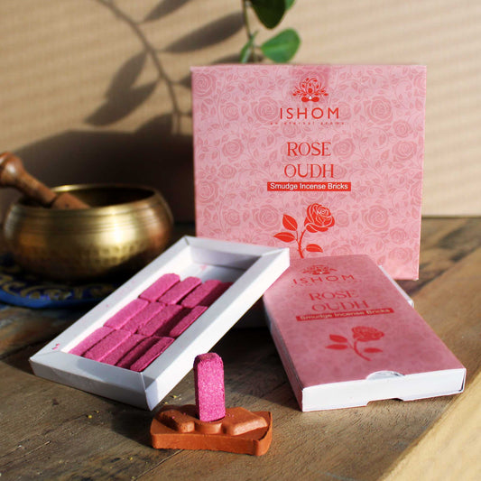 Pink incense bricks are displayed in an open box labeled "ISHOM Rose Oudh Smudge Incense Bricks." A single brick stands on a terracotta holder beside a brass bowl, on a wooden surface.