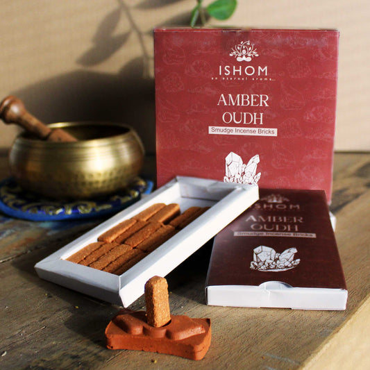 Incense bricks labeled "ISHOM AMBER OUDH" are arranged on wooden surface; a box and tray display them, while a brass bowl with a pestle rests in the background.