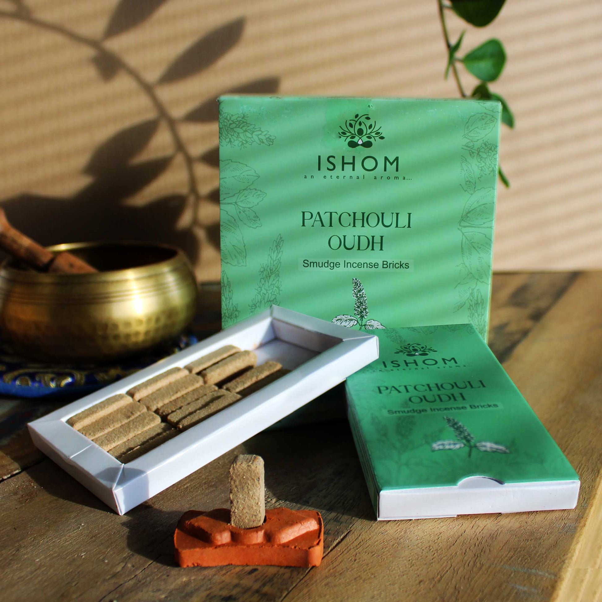 Incense bricks arranged in an open box on a wooden surface; labeled "ISHOM an eternal aroma... PATCHOULI OUDH Smudge Incense Bricks." A brass bowl and green leaves are nearby.