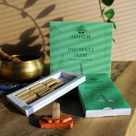Incense bricks arranged in an open box on a wooden surface; labeled "ISHOM an eternal aroma... PATCHOULI OUDH Smudge Incense Bricks." A brass bowl and green leaves are nearby.