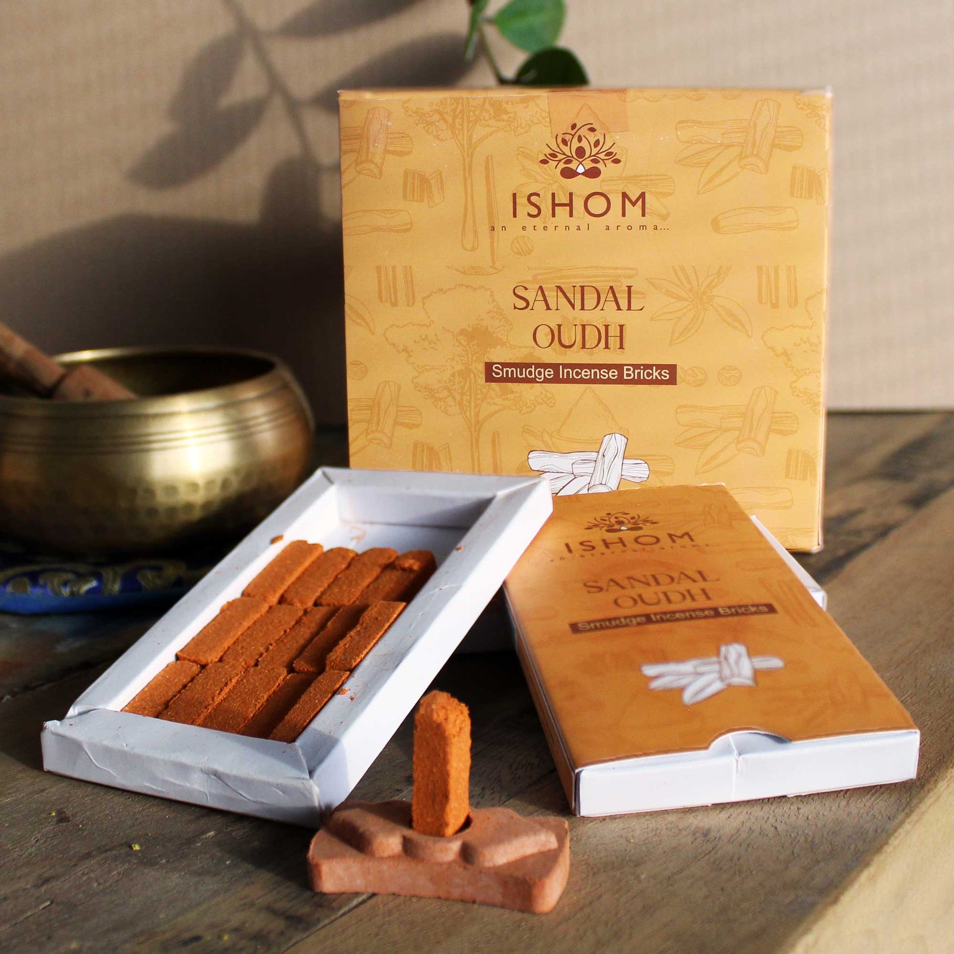 Incense bricks labeled "Sandal Oudh" are arranged beside an opened box. A lit brick stands on a small holder. Background features a brass bowl and a leafy plant. Text: "ISHOM an eternal aroma. SANDAL OUDH Smudge Incense Bricks."