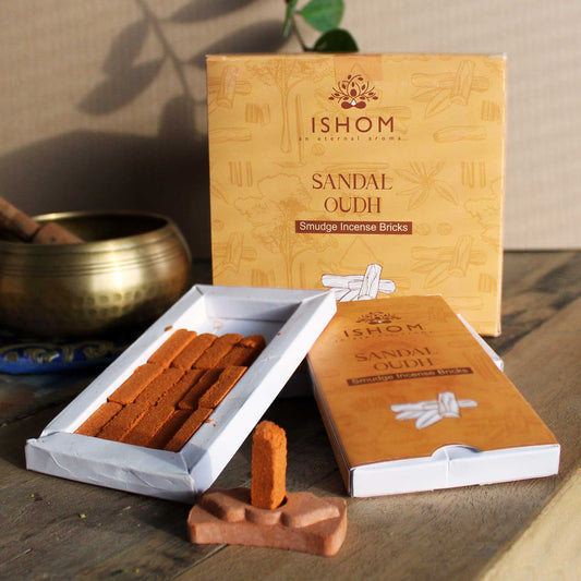 Incense bricks labeled "Sandal Oudh" are arranged beside an opened box. A lit brick stands on a small holder. Background features a brass bowl and a leafy plant. Text: "ISHOM an eternal aroma. SANDAL OUDH Smudge Incense Bricks."