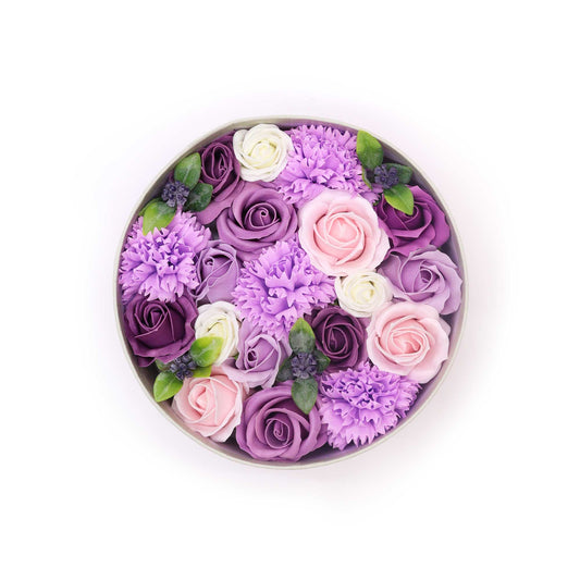 A circular box contains assorted artificial roses and carnations in shades of purple, pink, and white.  They are arranged densely, filling the container. The context is a plain white background.